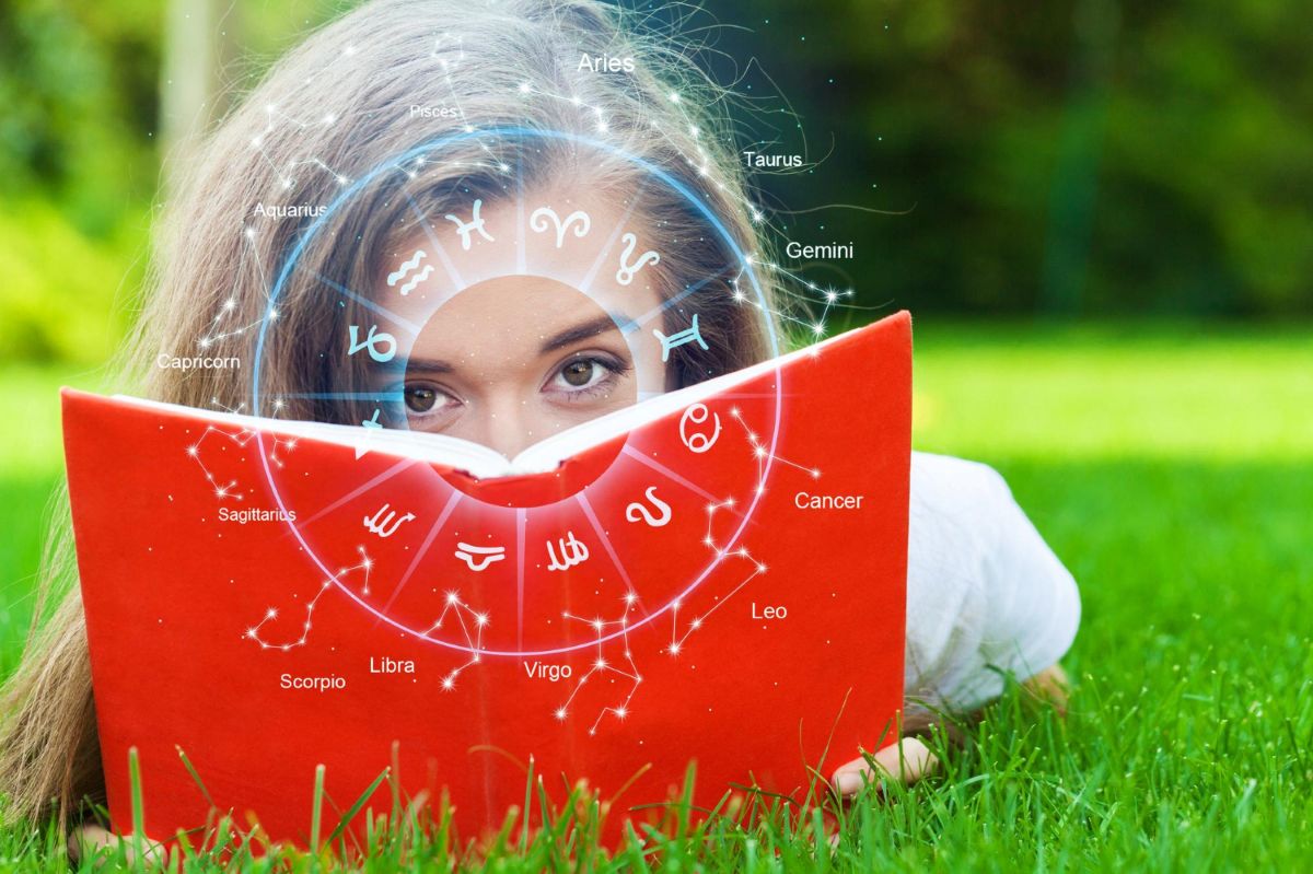 Moteris, laikanti raudoną knygą su Zodiako ženklais, žvelgia pro ją ant žalios žolės fono.