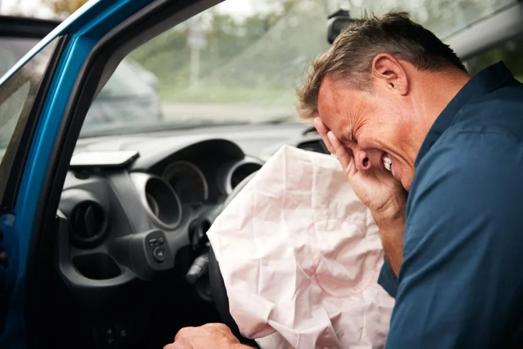 Žmogus sėdi automobilyje su išsiskleidusia oro pagalve, laikydamas ranką prie veido, vaizduoja emocinę įtampą.